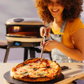 Eine Frau in einem gelben Hemd und Jeans-Overall lächelt, während sie mit dem Gozney Utility runder Pizzawender mit ergonomischem Design eine frisch gebackene Pizza auf einem Paddel schneidet. Im Hintergrund ist ein moderner Pizzaofen zu sehen.