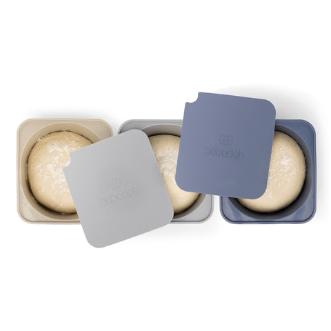 Drei Babadoh Silikon BIG Pizzateig und Zutatenbehälter 3er-Set (Multi Blue) von Babadoh, in den Farben beige, hellgrau und blau, halten jeweils eine Teigkugel, deren Deckel leicht geöffnet ist, um den Inhalt zu zeigen.