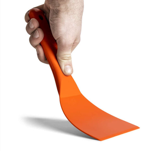 Eine Hand hält einen orangefarbenen FUMOSA flexiblen Teigspachtel aus Kunststoff mit langem Griff und flacher, abgeschrägter Kante. Die Hand umklammert den ergonomischen Griff fest und der Schaber ist nach unten abgewinkelt, bereit zum Gebrauch – ideal für Pizza-Enthusiasten.
