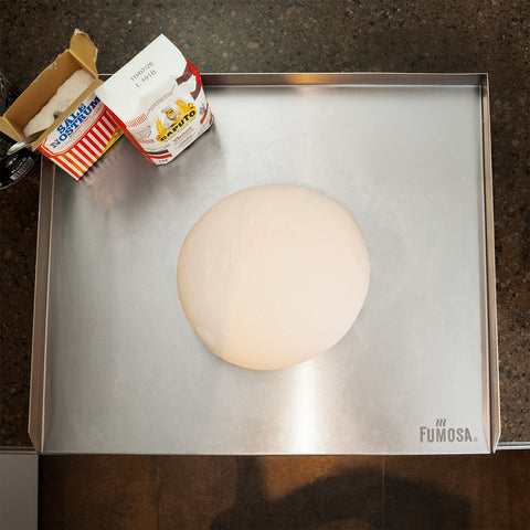 Eine runde Teigkugel liegt auf der FUMOSA IMPASTO Edelstahl Arbeitsplatte inkl. Silikon Antirutschmatte (60x50) von Fumosa, mit Backzutaten in der Nähe, bereit für die weitere Zubereitung.