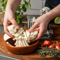 Eine Person legt frisch geschnittenen Mozzarella in eine braune Schüssel, im Hintergrund ein FUMOSA Profi Mozzarellaschneider. Um die Schüssel herum stehen frische Kräuter, Tomaten und eine Basilikumpflanze, alles auf einer Holzoberfläche in einer Küchenumgebung.