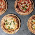 Vier Holzofenpizzas mit verschiedenen Belägen sind auf einem Fumosa FUMOSA Pizza-Servierteller PIATTO aus Bambus, 2er-Set auf einer dunkelgrauen Arbeitsplatte angerichtet. Eine Fumosa-Pizza hat Tomaten und Basilikum, eine andere Käse und Basilikum, die dritte hat Pilze und Basilikum und die vierte hat Kirschtomaten und Basilikum.