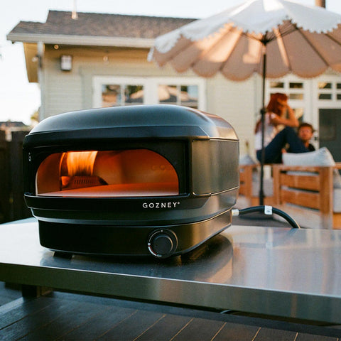 Ein Gozney Arc Lite Gas Pizzaofen von Gozney steht auf einem Tisch im Freien, die Flammen glühen in seiner 500 °C heißen Gaskammer, während zwei Personen auf einer Terrasse unter einem großen Sonnenschirm vor einem Haus entspannen.