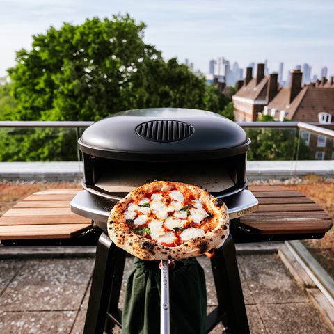 Eine frisch gebackene Pizza mit geschmolzenem Käse und Kräutern wird aus dem eleganten Gozney Ofensims & Frontablage für Arc XL-Außenofen auf der Dachterrasse gehoben. Die Skyline der Stadt und die Bäume bilden eine malerische Kulisse unter dem klaren Himmel und verstärken das ultimative Gozney-Erlebnis.