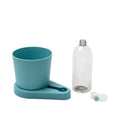 Ein Wasserspenderset aus Kunststoff für Haustiere, bestehend aus einem blaugrünen Wassernapf auf einem Sockel, einer leeren durchsichtigen Plastikflasche und einem weißen Deckel mit einer Düse. Die Flasche ist so konzipiert, dass sie in den Ständer umgedreht werden kann, um Wasser in den Napf zu geben, ähnlich wie unser greevi Kräuterheld selbstbewässernder Kräuter-Pflanztopf (GREEVI) für frische Kräuter von Grevi.