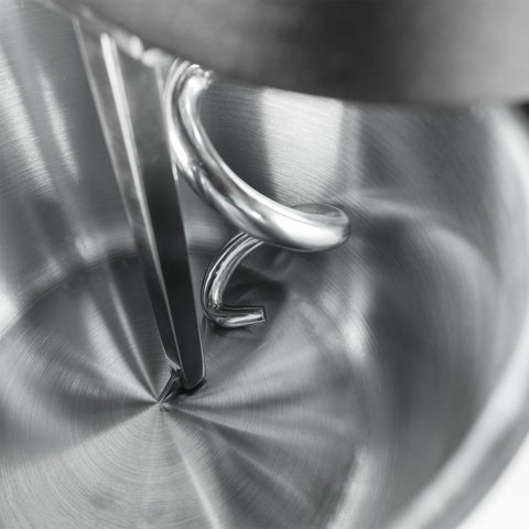 Nahaufnahme einer KernKraft KnetKraft 5 Spiral-Knetmaschine mit einem Edelstahlkessel, wobei der Knethaken und die Flachrühreraufsätze hervorgehoben werden, die das Licht von den glänzenden Metalloberflächen reflektieren.