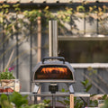 Der Ooni Karu 2 Pro Pizzaofen von Ooni mit Edelstahlkamin und ClearView-Glastür steht auf einem Tisch zwischen Pflanzen und Pizzazutaten, im Inneren glüht ein Feuer, während im Hintergrund ein Gebäude aufragt.