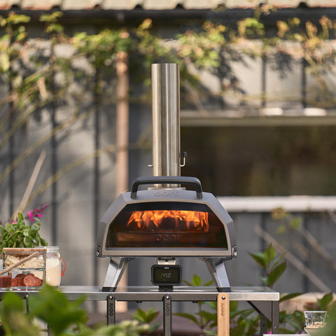 Der Ooni Karu 2 Pro Pizzaofen von Ooni mit Edelstahlkamin und ClearView-Glastür steht auf einem Tisch zwischen Pflanzen und Pizzazutaten, im Inneren glüht ein Feuer, während im Hintergrund ein Gebäude aufragt.