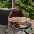Eine Hand schiebt eine ungebackene Pizza auf einer Holzschaufel in den schwarzen Ooni Karu 2 Pro Pizzaofen von Ooni, im Hintergrund ist grünes Laub verschwommen.