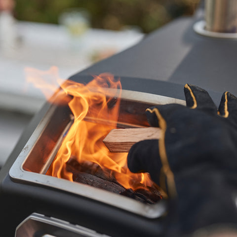 Eine behandschuhte Hand legt Holz in die brennende Flamme eines Ooni Karu 2 Pro Pizzaofens von Ooni. Leuchtend orange Flammen flackern hinter der ClearView-Glastür, während der Hintergrund unscharf erscheint.