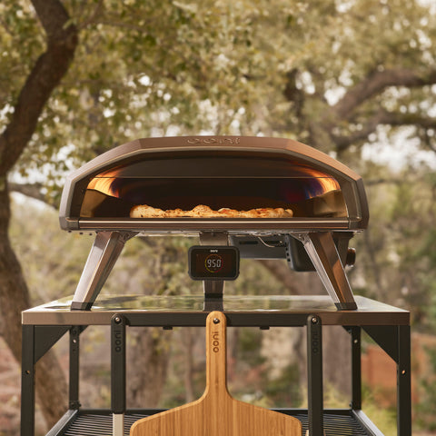Eine Pizza wird in einem Ooni Koda 2 Pro Gas-Pizzaofen von Ooni auf einem Metalltisch gebacken, darunter befindet sich ein Pizzaschieber aus Holz und im Hintergrund ist verschwommenes Grün zu sehen.