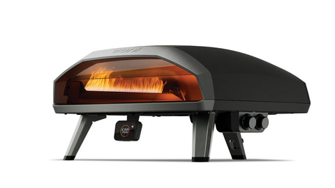 Un élégant four à pizza d'extérieur noir au design moderne. Le four à pizza à gaz Ooni Koda 2 Max - 61 cm d'Ooni repose sur quatre pieds coudés et a une face avant ouverte qui montre une flamme orange à l'intérieur. Équipé de deux zones de cuisson et d'un indicateur de température sous la porte du four, il est clair que ce four à pizza à gaz est en cours d'utilisation.