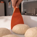 Eine Hand hält einen orangefarbenen FUMOSA flexibler Teigspachtel von Fumosa und hebt eine runde Teigkugel aus einem weißen Behälter. Im Vordergrund sind weitere Teigkugeln zu sehen, bereit für Pizza-Enthusiasten, die sie in einer Küchenumgebung mit unscharfen Hintergrundelementen verwenden können.