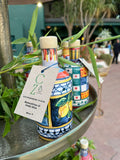 Eine bunt bemalte, handgefertigte Keramikflasche mit Cunzatillu Olivenöl mit Etikett hängt auf einem Display zwischen grünem Laub. Auf dem Etikett steht „Olio Extra Vergine di Oliva“. Andere ähnlich dekorierte Flaschen sind im Hintergrund zu sehen, draußen in einem sonnigen Bereich mit Grün und Menschen, die sizilianische Tradition zur Schau stellen.