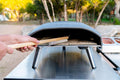 Eine Person verwendet eine Ooni Ooni Pizzaofenbürste mit Metallborsten, um die Steinoberfläche im Inneren eines tragbaren Outdoor-Pizzaofens zu reinigen. Der graue Ofen steht auf einem Metalltisch, mit Bäumen und Landschaft im unscharfen Hintergrund.
