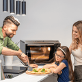 Ein Mann schneidet mit einem Pizzaschneider eine Pizza auf einer Arbeitsplatte. Ein junges Mädchen und eine Frau stehen neben ihm und schauen lächelnd zu. Im Hintergrund ist der Effeuno Pizzaofen EffeUno P134HA 509°C zu sehen, in dem sich eine Pizza befindet, die unter der modernen Hängelampe über ihnen die höchsten Backtemperaturen erreicht.