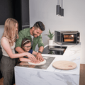 Eine Frau, ein Mann und ein junges Mädchen bereiten gemeinsam Teig auf einer Küchentheke aus weißem Marmor zu. Sie lächeln und konzentrieren sich darauf, den Teig für Backen von Pizza Napoletana zu formen. Im Hintergrund sind moderne Küchengeräte zu sehen, darunter ein Effeuno Pizzaofen EffeUno P134HA 509°C und ein Schneidebrett.