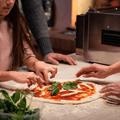 Ein Kind und zwei Erwachsene bereiten in einer Küche eine Pizza zu. Das Kind legt grüne Basilikumblätter auf eine Pizza mit Tomatensauce und Käse, während die Hände der Erwachsenen geschickt den Belag anordnen. Im Vordergrund sind frische Basilikumblätter zu sehen, bereit, mit dem Effeuno Pizzaofen EffeUno P134H 509°C eine authentische Pizza Napoletana zuzubereiten.