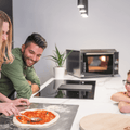 Eine Frau verteilt mit einem Löffel Tomatensoße auf Pizzateig, während ein Mann und ein Kind lächelnd zuschauen. Sie befinden sich in einer modernen Küche mit weißer Arbeitsplatte, einem Backofen im Hintergrund und einer Topfpflanze in der Nähe. Ein weiterer Pizzateig liegt auf einem Holzbrett auf der Arbeitsplatte, bereit zum Backen im Pizzaofen EffeUno P134HA 509°C von Effeuno.