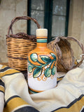 Eine dekorative handgefertigte Keramikflasche mit Cunzatillu Olivenöl, bemalt mit einem Oliven- und Blättermotiv, das die sizilianische Tradition verkörpert, steht auf einem gestreiften Tuch. Hinter der Cunzatillu-Flasche befinden sich zwei geflochtene Körbe. Die Szene scheint sich in einem Innenbereich vor einem Fenster mit zwei Scheiben abzuspielen.