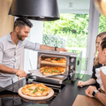 Ein Mann nimmt eine frisch gebackene Pizza aus einem Effeuno Pizzaofen Effeuno P234H 509°C mit 2 Backkammern in einer hellen, modernen Küche. Zwei Frauen stehen an der Seite, schauen zu und lächeln. Eine weitere Pizza auf einem Biscotto-Stein steht bereits auf der Theke, garniert mit Basilikumblättern. Die Küche öffnet sich zu einem grünen, üppigen Hinterhof.